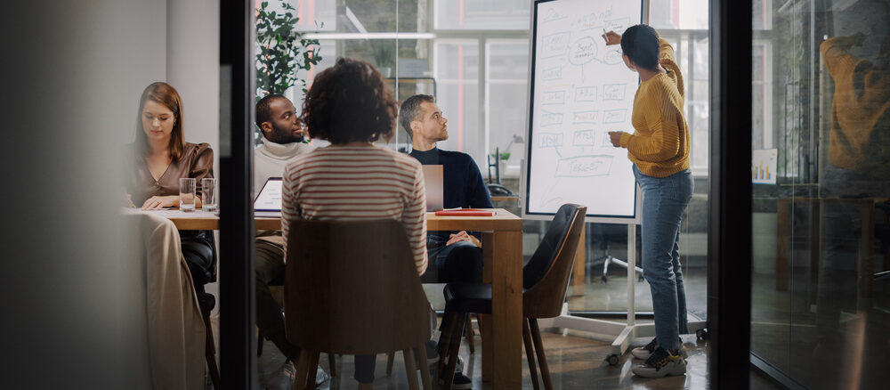 Project Manager Makes a Presentation for a Young Diverse Creative Team in Meeting Room in an Agency. Colleagues Sit Behind Conference Table and Discuss Business Development, User Interface and Design