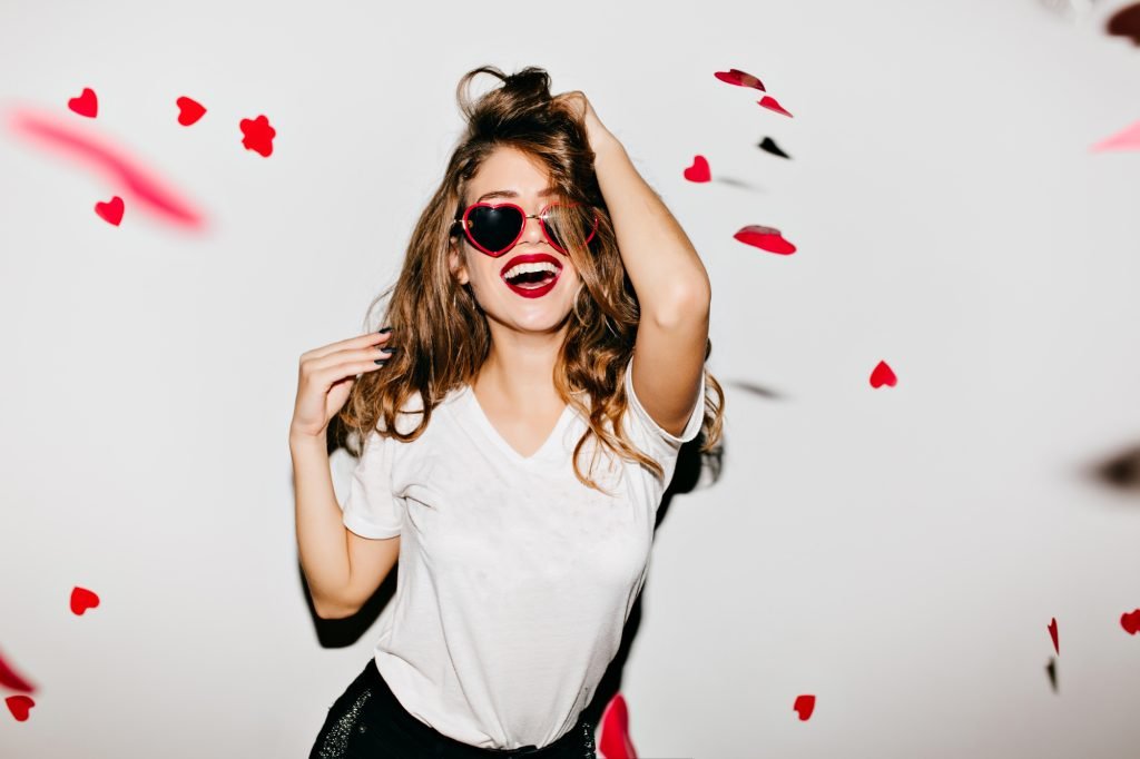 Indoor portrait of amazing caucasian female model in trendy t-shirt touching her long shiny hair. Laughing refined woman in sunglasses having fun with red confetti.