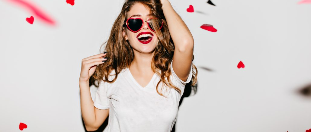 Indoor portrait of amazing caucasian female model in trendy t-shirt touching her long shiny hair. Laughing refined woman in sunglasses having fun with red confetti.