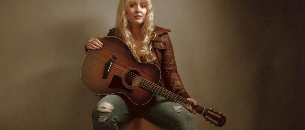 young blond woman sitting on a chair wearing blue jeans and brown jacket playing the guitar