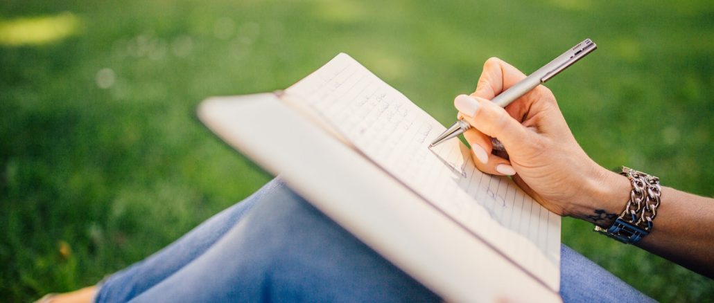 women journaling sitting on green grass holding a pen