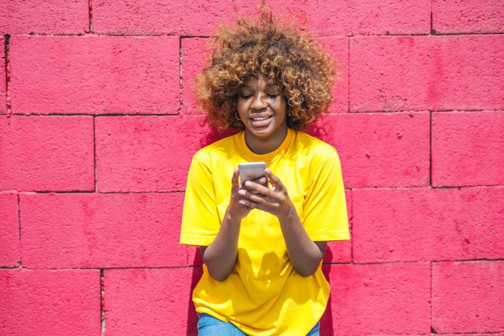 woman wearing a yellow t shirt, against pink wall, looking at the phone