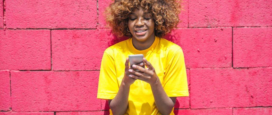 woman wearing a yellow t shirt, against pink wall, looking at the phone