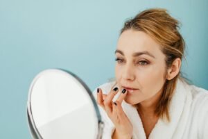 A woman in a white robe applying skincare cream to her lips while looking into a mirror, symbolizing a daily beauty and self-care routine for healthy skin. Secrets of Health and Longevity