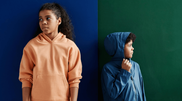 Two children stand in front of a two-toned background, with the left half in deep blue and the right in forest green. The child on the left wears a peach hoodie and gazes to the side, while the child on the right, wearing a blue hooded jacket, looks in the opposite direction. The scene conveys a casual yet thoughtful mood.