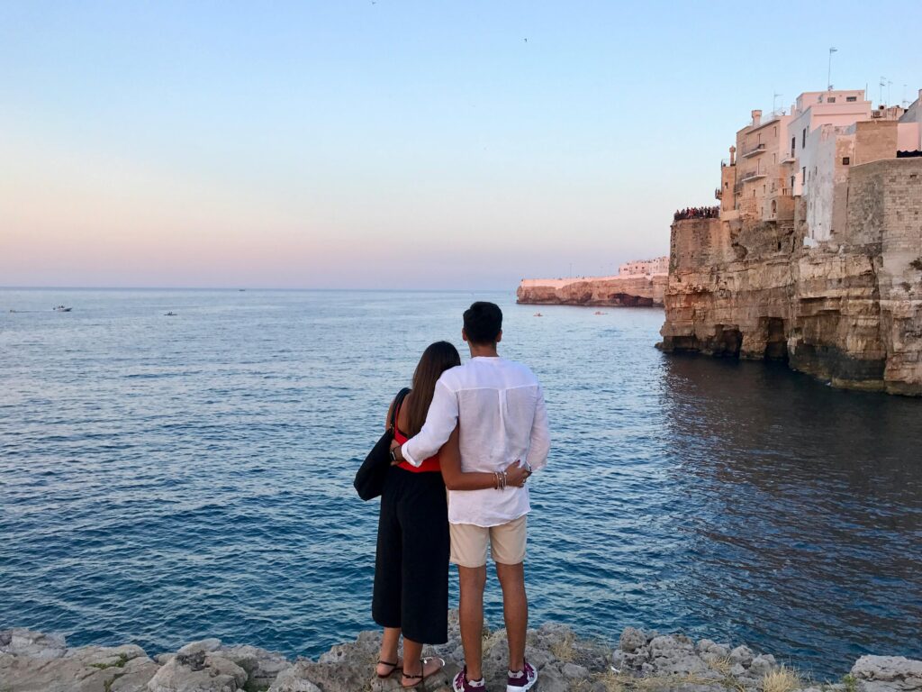 man-in-white-dress-shirt-standing-beside-the-woman-in-black-and-red-dress-while-watching-the-blue-calm-water-near-brown-concrete-buildings-under-white-and-blue-sky-at-daytime