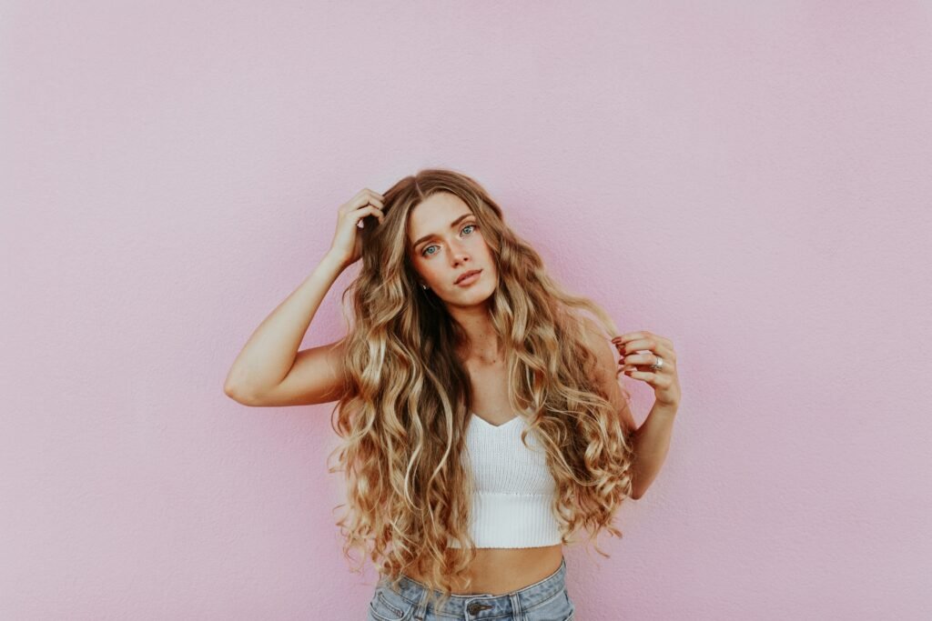 Elegant woman with long wavy blonde hair in front of a pink background, representing holistic beauty and anti-aging secrets