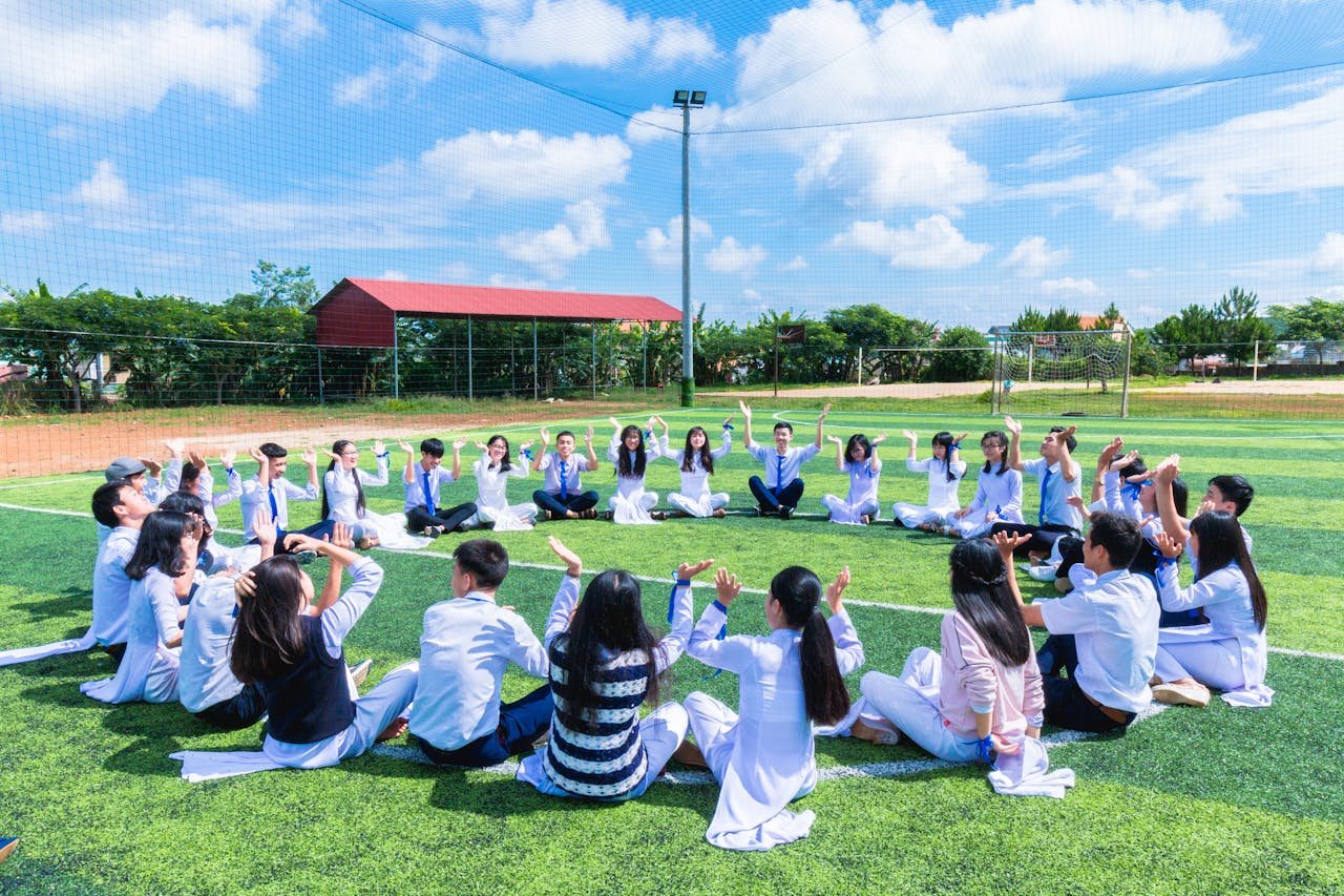 A group of students sitting in a circle on a grassy field during a collaborative outdoor activity, symbolizing teamwork and efficiency—concepts central to running a school like a business. The bright blue sky and organized setting reflect structure and focus