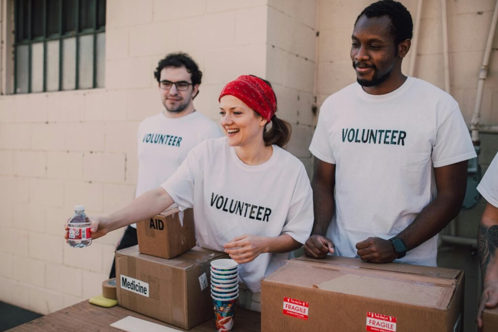 Group of volunteers in white t-shirts distributing aid, including water bottles and labeled boxes of medicine and supplies, smiling and working together outdoors