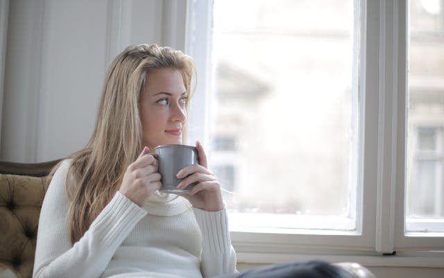 woman wearing white sweater - positive mindset - woman holding a cup looking at her window with a smile - pump it up magazine