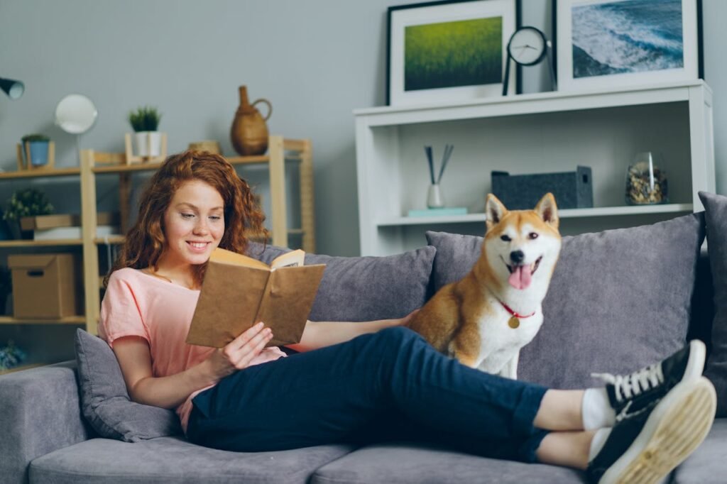 A smiling woman reading a book on a cozy couch with her happy Shiba Inu dog beside her.