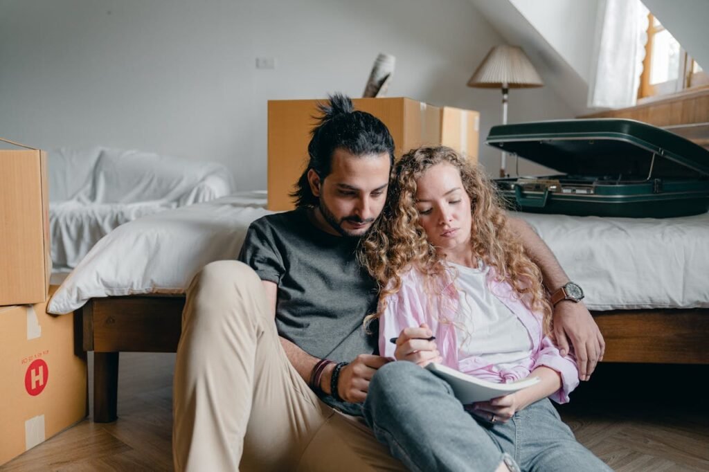 Free Thoughtful male and female in casual wear sitting near bed among boxes together and taking notes while moving house Stock Photo